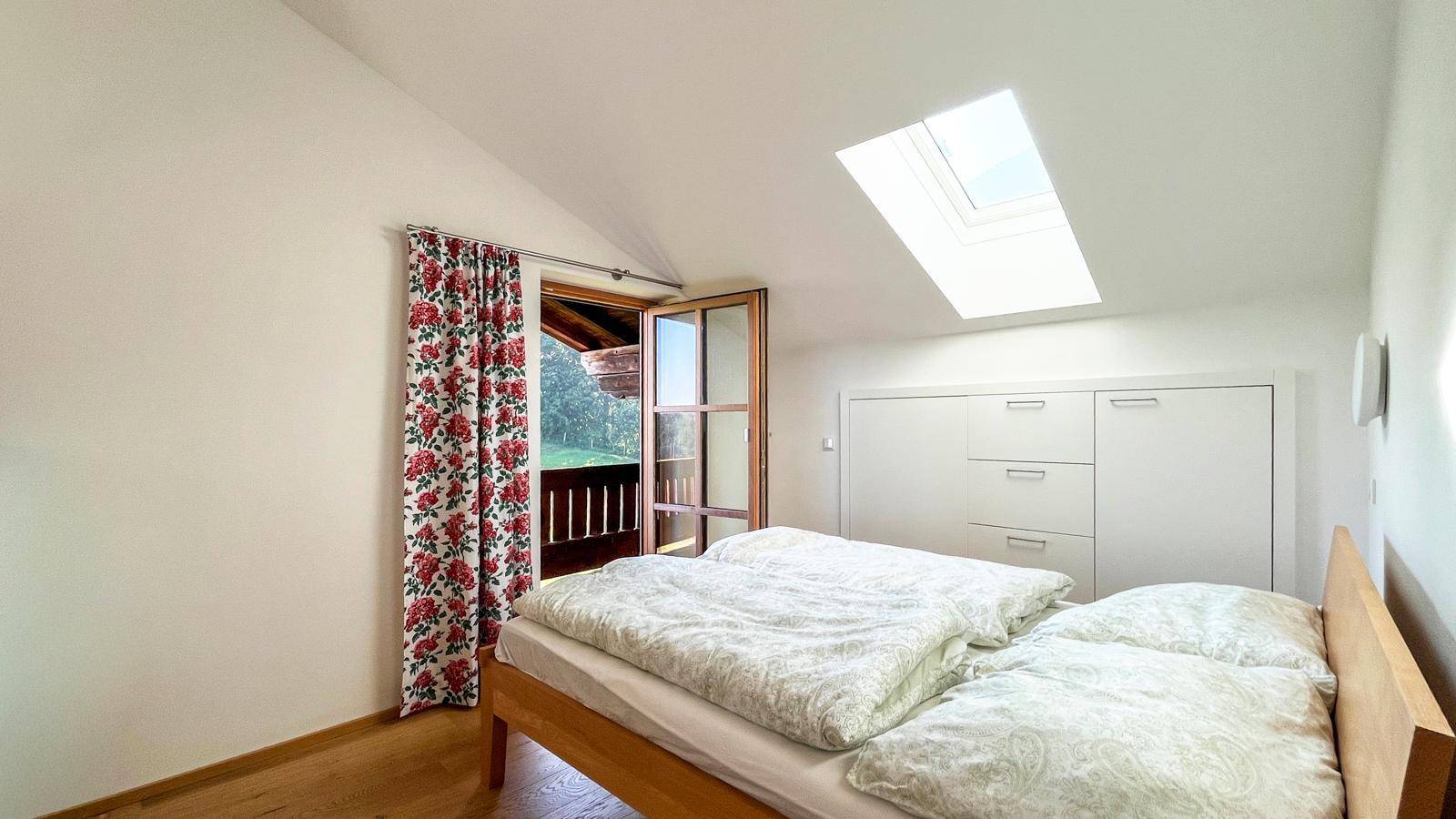 Schlafzimmer mit Holzbett, Zugang zum Balkon und Dachfenster, das viel natürliches Licht hereinlässt.