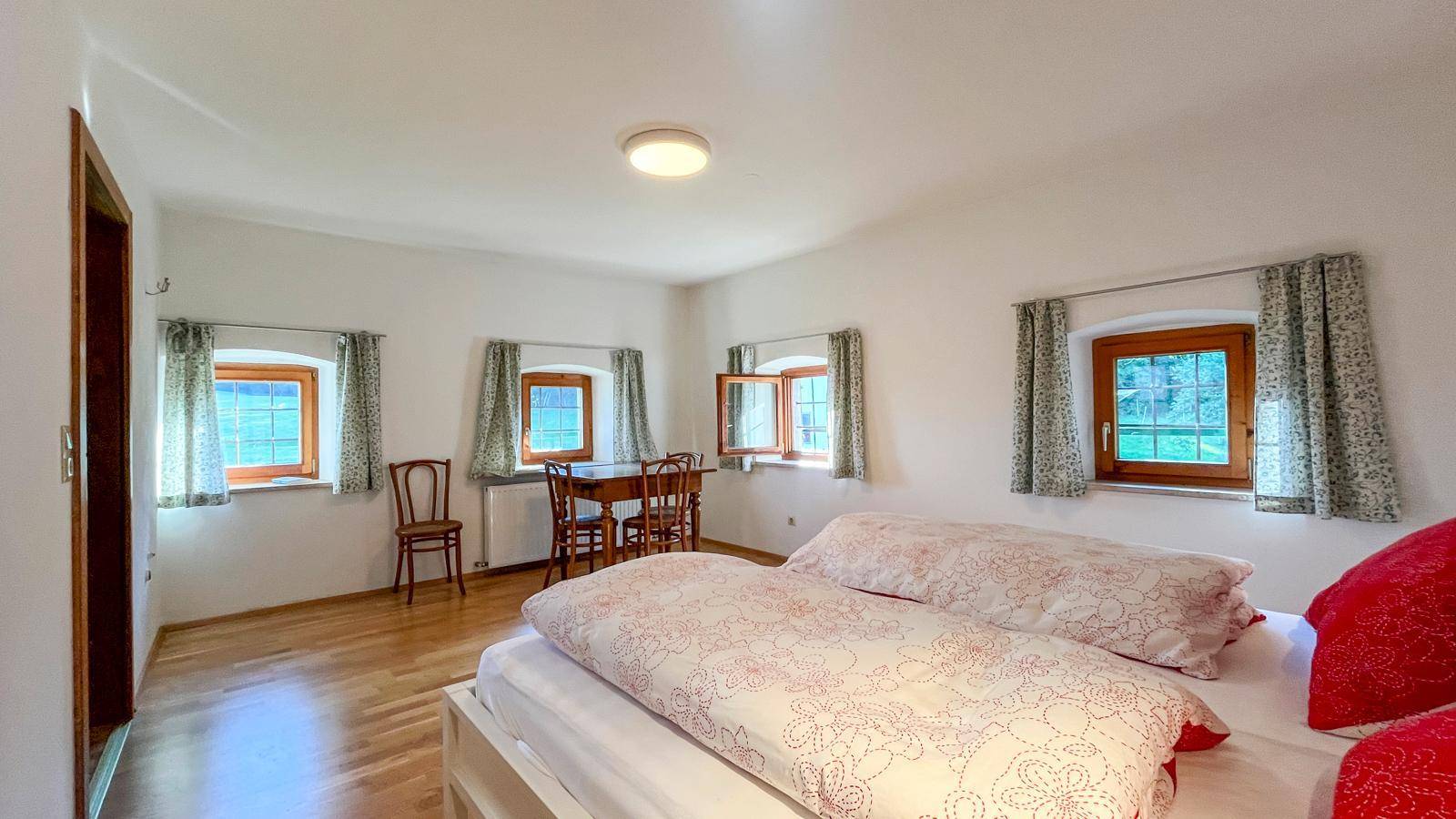 Helles Schlafzimmer mit Holzboden, mehreren Fenstern und einem kleinen Tischbereich, bietet viel Tageslicht.