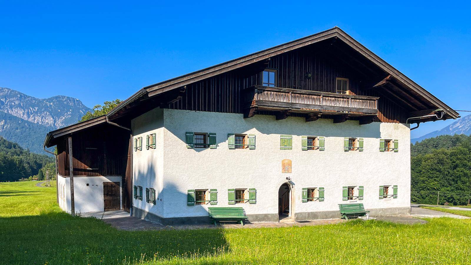 Nahaufnahme des traditionellen Bauernhauses mit Holzbalkon und grünen Fensterläden, umgeben von grünen Wiesen.
