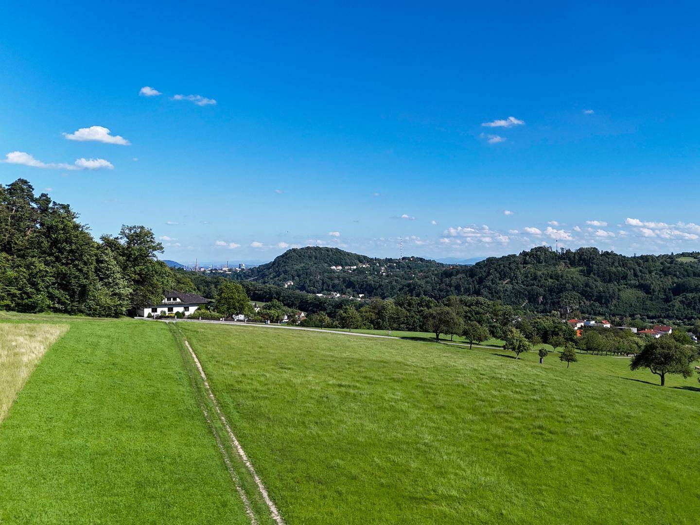 Weite grüne Wiesenfläche mit einem Haus am Horizont und bewaldeten Hügeln unter blauem Himmel.