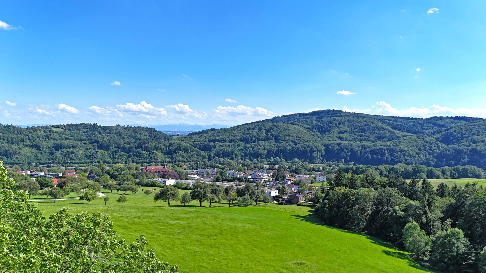 Panoramablick auf ein Dorf in einem grünen Tal, umgeben von bewaldeten Hügeln.