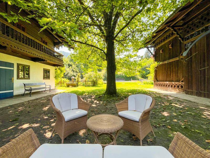 Gemütlicher Sitzbereich im Garten unter einem großen Baum, umgeben von traditionellen Gebäuden.