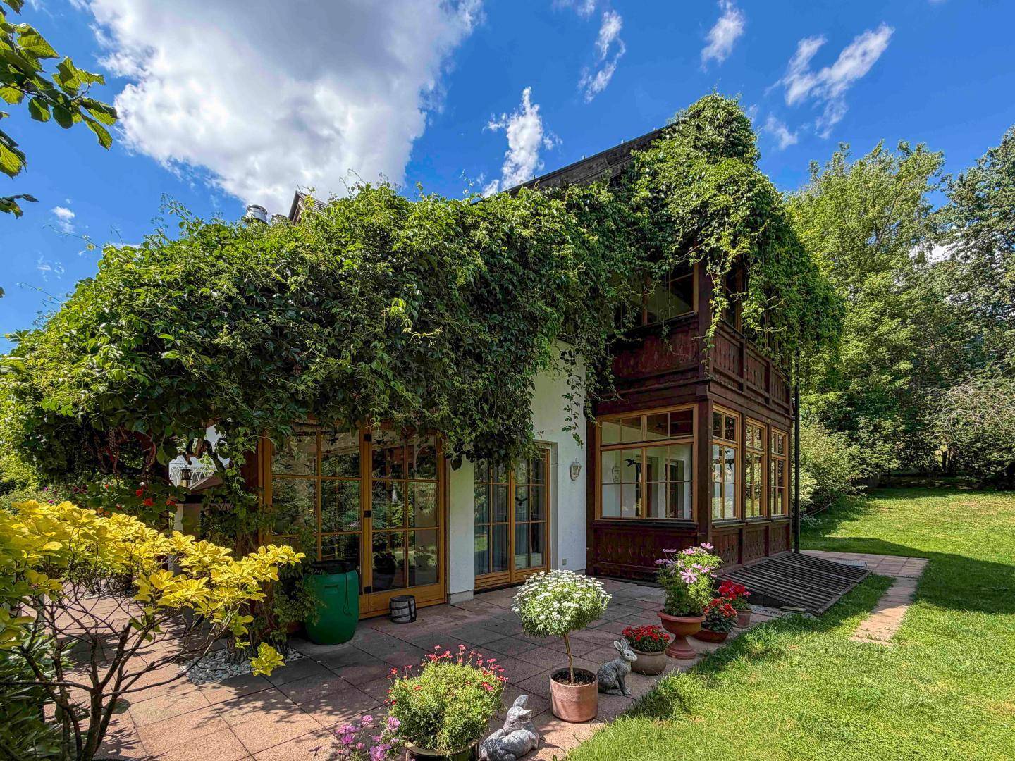 Charmantes Haus mit bewachsener Fassade und blühenden Topfpflanzen auf der Terrasse, umgeben von Grün.