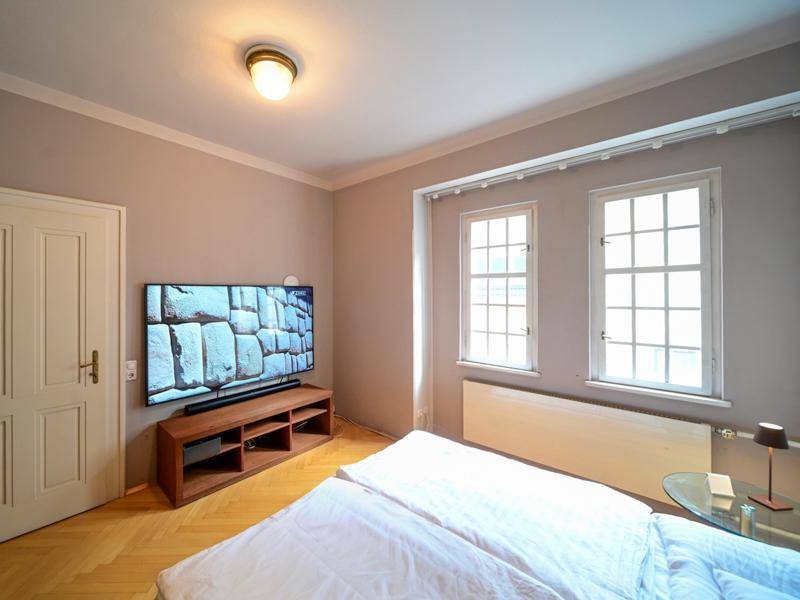 Helles Schlafzimmer mit Doppelbett, Fernseher, Holzboden und zwei Fenstern.