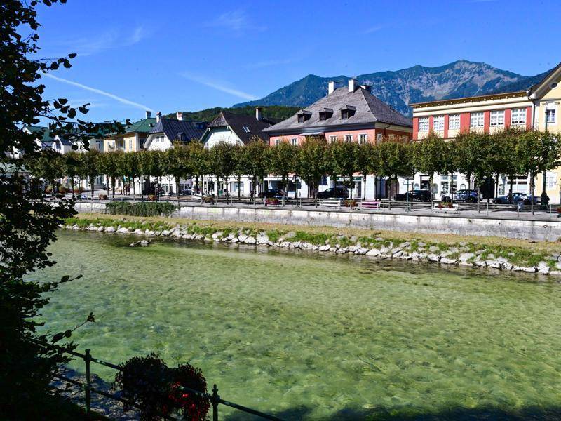 Historische Gebäude entlang eines Flusses mit Baumallee und Bergen im Hintergrund.