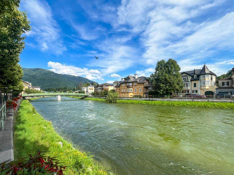 Flusslandschaft mit Brücke, Gebäuden und Bergen unter einem weiten blauen Himmel.