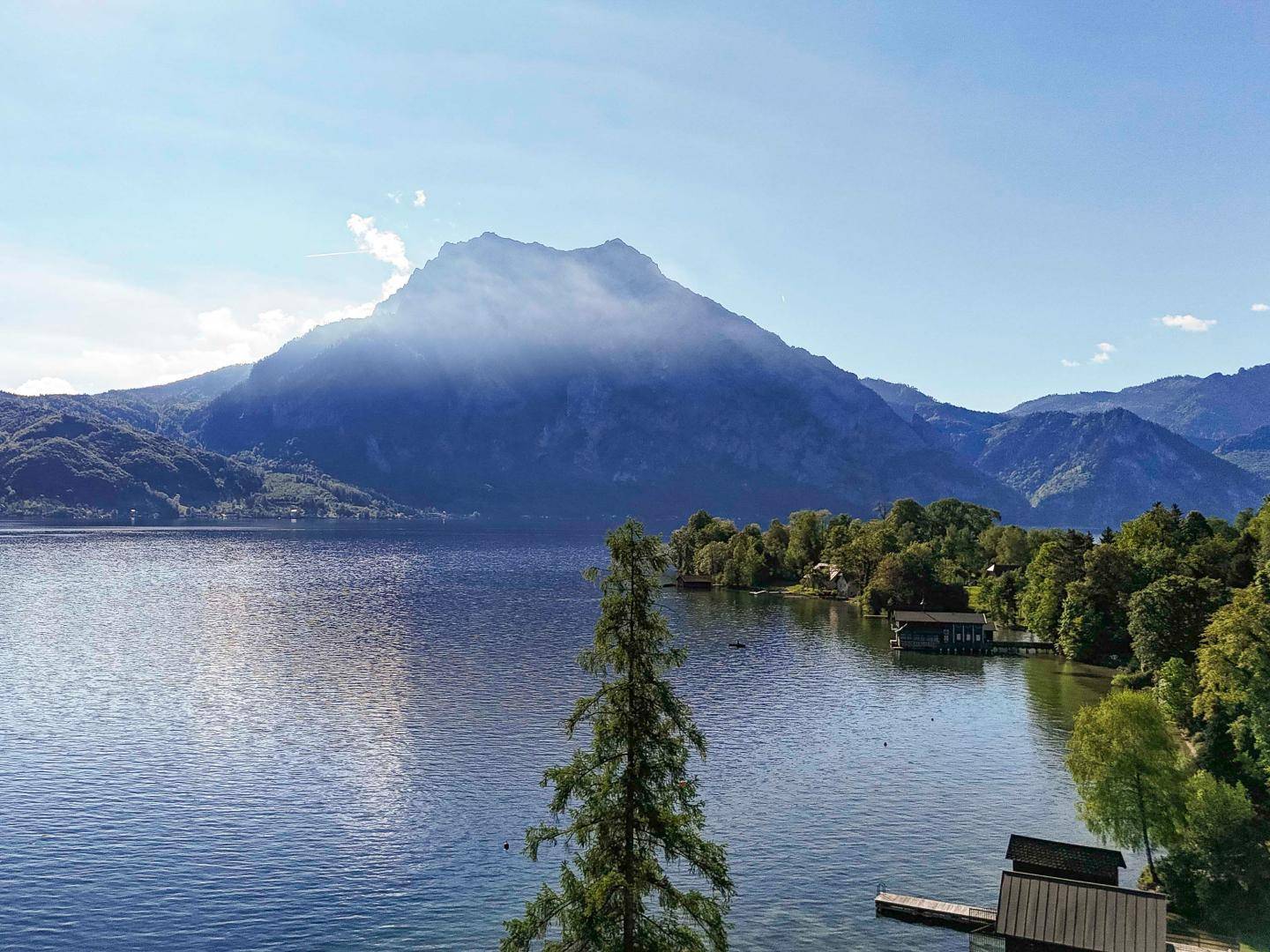 Malerischer Blick auf den See mit einem nebelverhangenen Berg im Hintergrund und Bootshäusern am Ufer.
