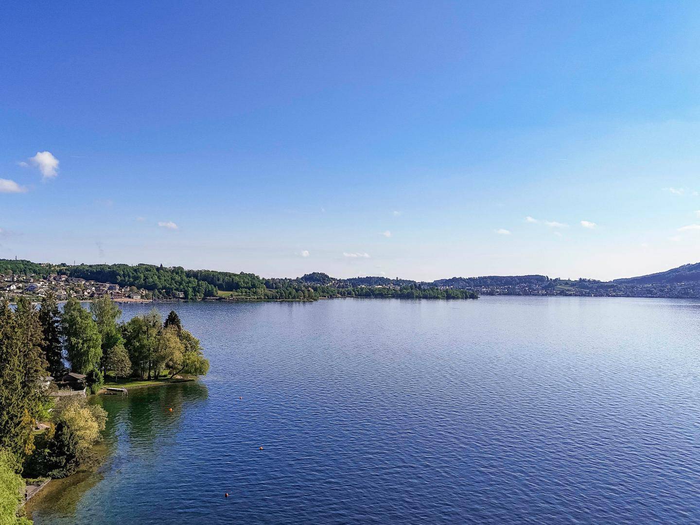 Weitläufige Luftaufnahme eines großen Sees, umgeben von grünen Ufern und Siedlungen am Horizont.