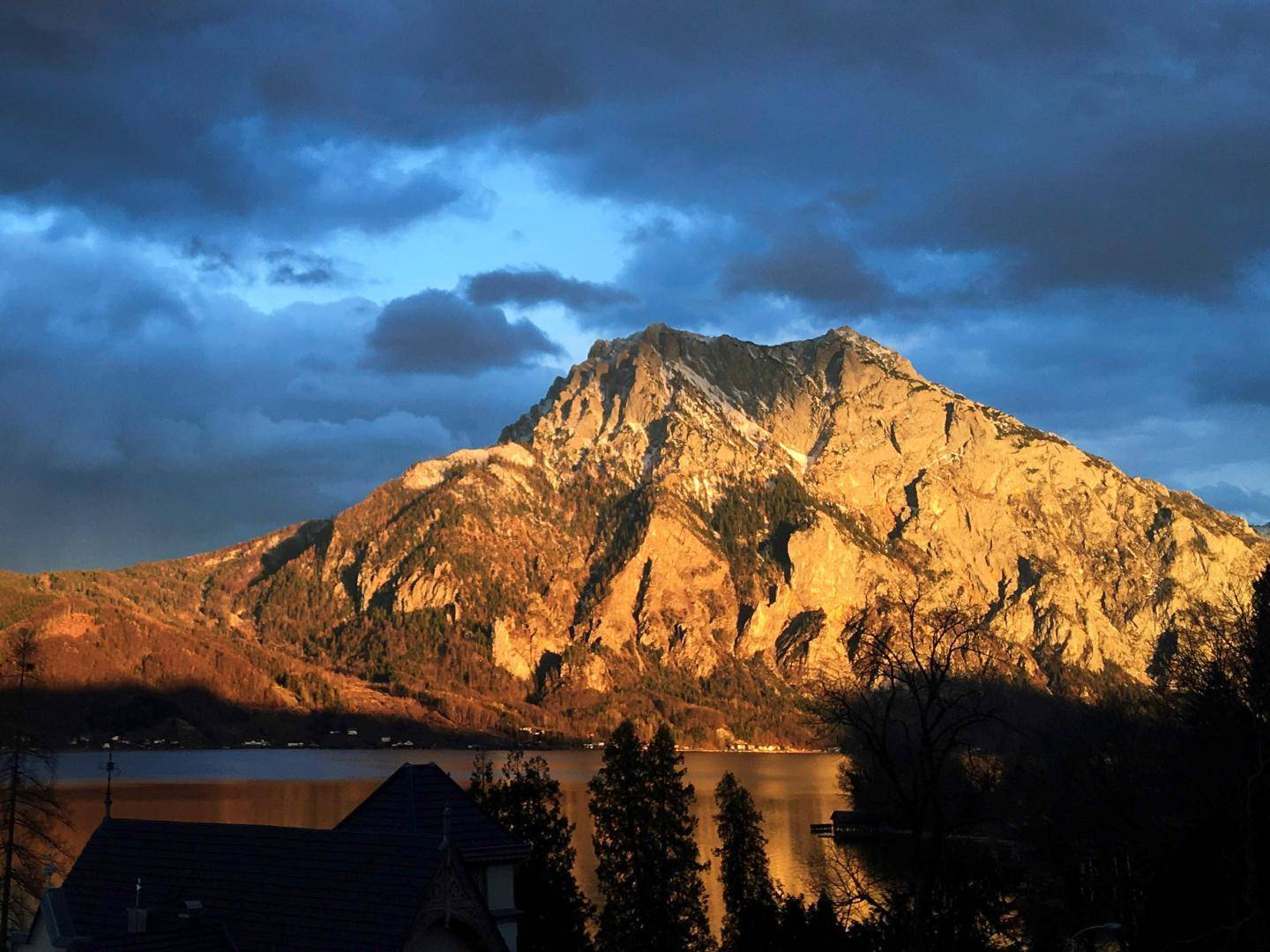 Majestätischer Berg im goldenen Abendlicht unter dramatischem Himmel mit See und Hausdach im Vordergrund.