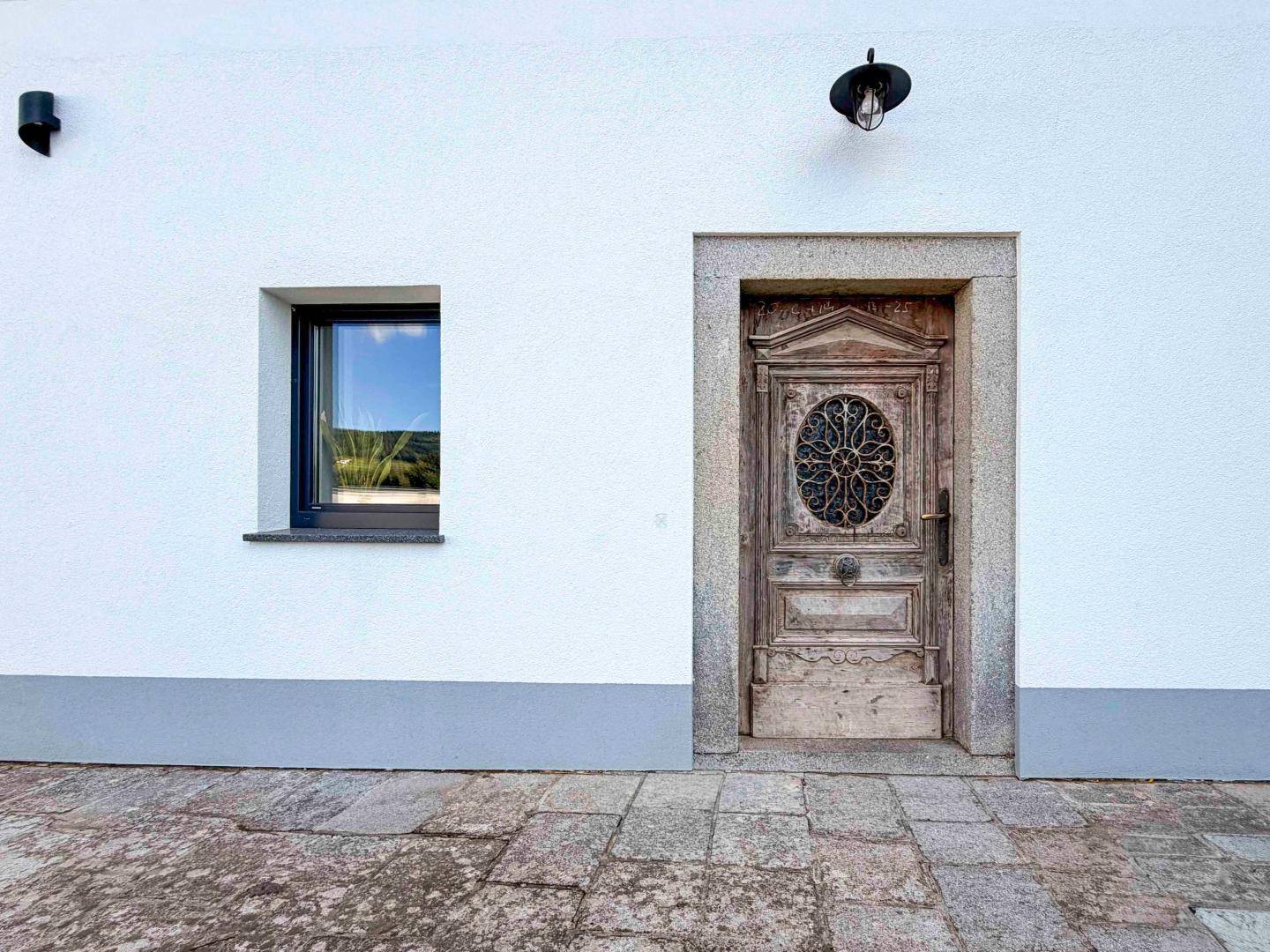 Historische Holztür mit Steinrahmen in einer weiß verputzten Fassade mit kleinerem Fenster.