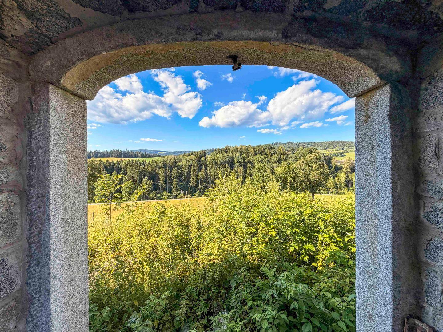Malerischer Ausblick durch einen Steinbogen auf grüne Wälder und Felder unter blauem Himmel.