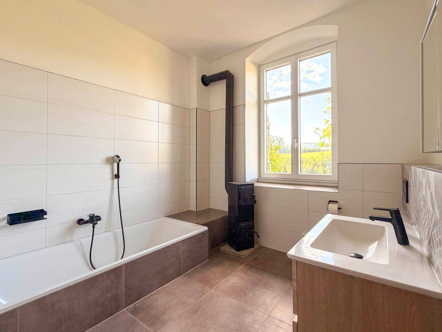 Modernes Badezimmer mit Badewanne, Waschbecken, Kaminofen und Fenster mit Weitblick.