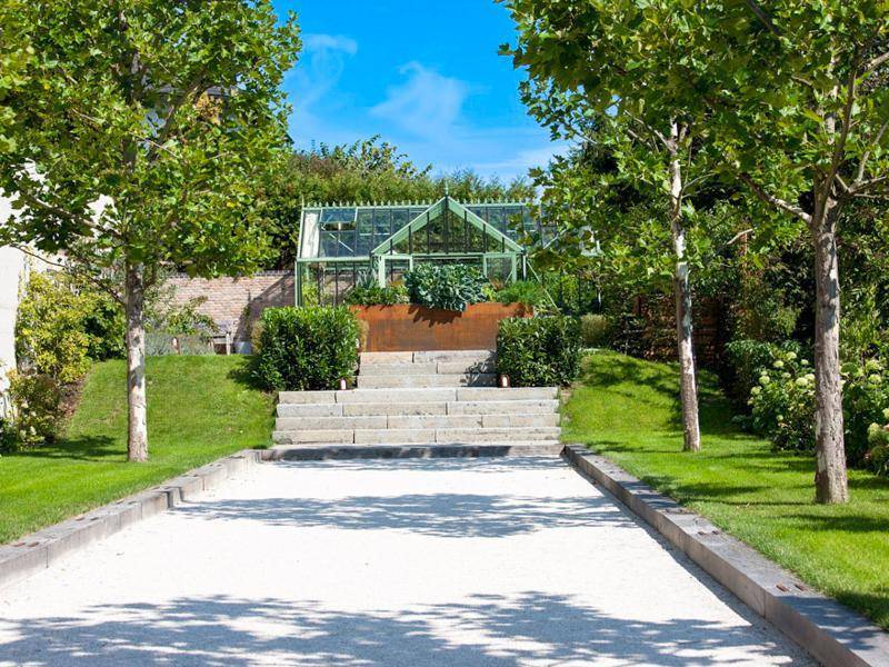 Gepflegter Gartenweg führt zu einem charmanten Gewächshaus, umgeben von Rasen und Bäumen.