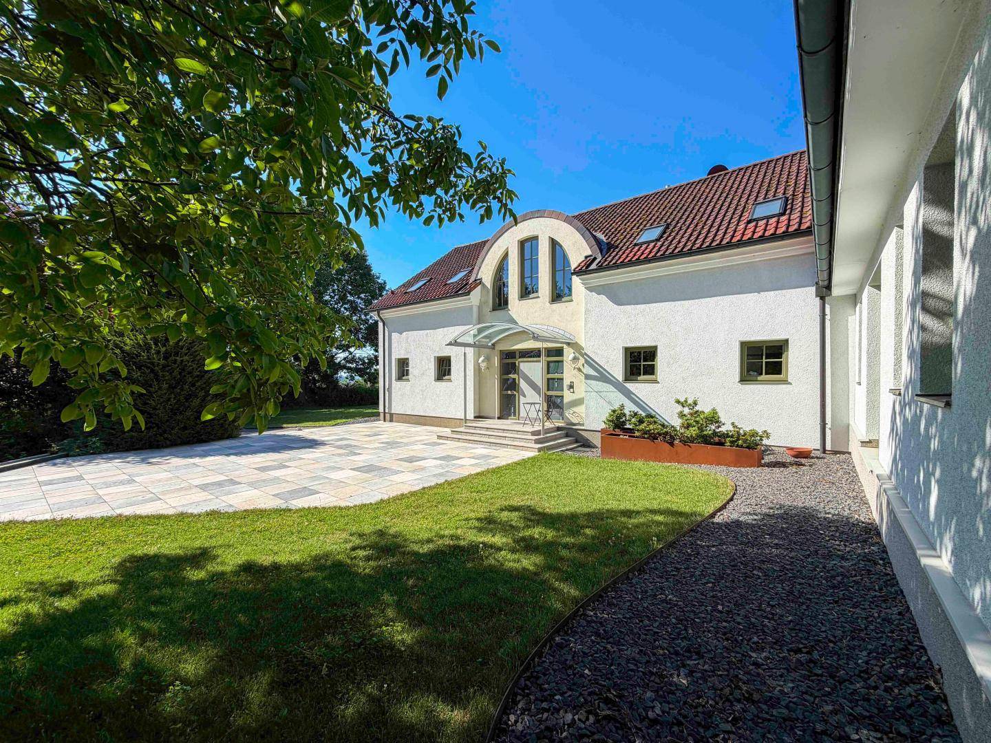 Seitlicher Eingangsbereich mit gepflasterter Terrasse und Kiesweg, umgeben von Grün.