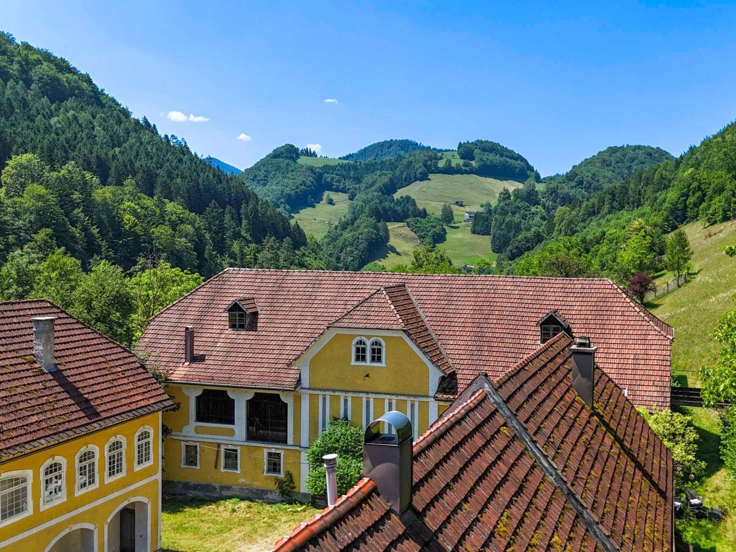 Luftaufnahme des Anwesens mit roten Dächern und gelben Fassaden, eingebettet in eine grüne Hügellandschaft.