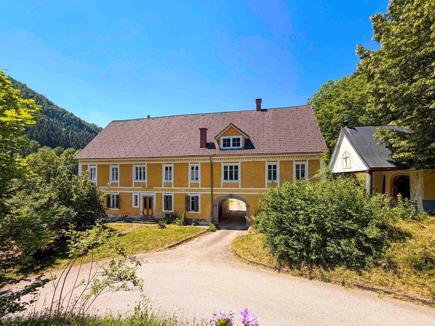 Vorderansicht des Hauptgebäudes mit gelber Fassade, rotem Dach und einem Torbogen, umgeben von Bäumen.