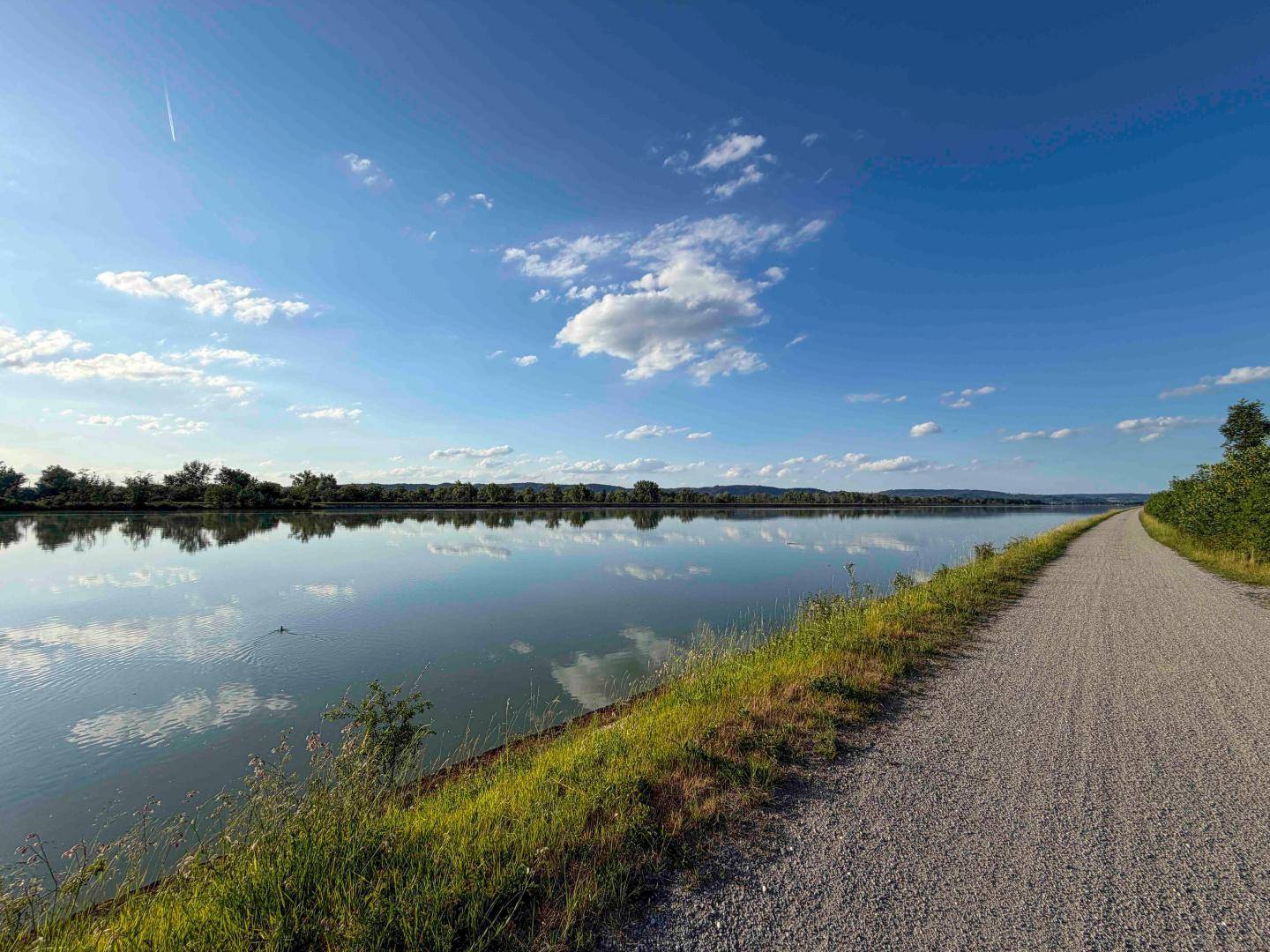 Ruhiger See oder Fluss mit einem Uferweg und bewaldeten Ufern unter blauem Himmel.