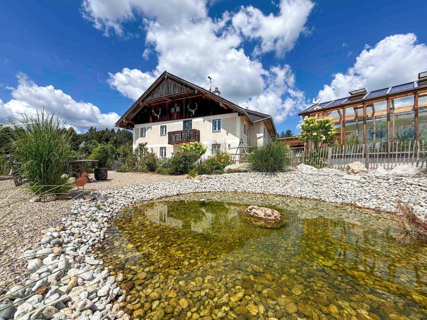 Landhaus mit idyllischem Teich im Vorgarten und umgebender Natur unter blauem Himmel.