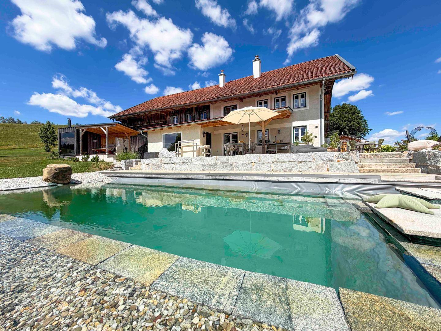Landhaus mit großem Naturpool und weitläufiger Terrasse unter strahlend blauem Himmel.
