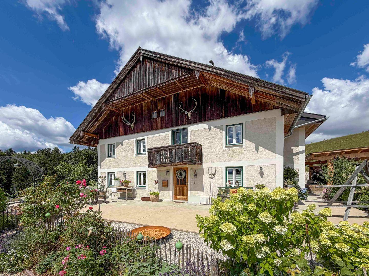 Charmante Fassade eines Landhauses mit Holzbalkon, Hirschgeweihen und blühendem Vorgarten.