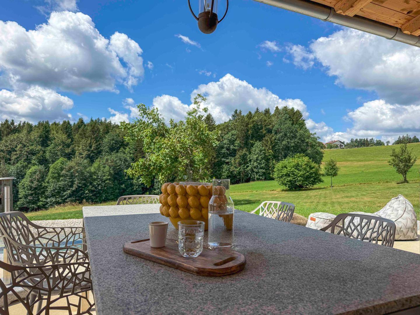 Detailaufnahme eines Terrassentisches mit Getränken und idyllischem Blick ins Grüne unter blauem Himmel.