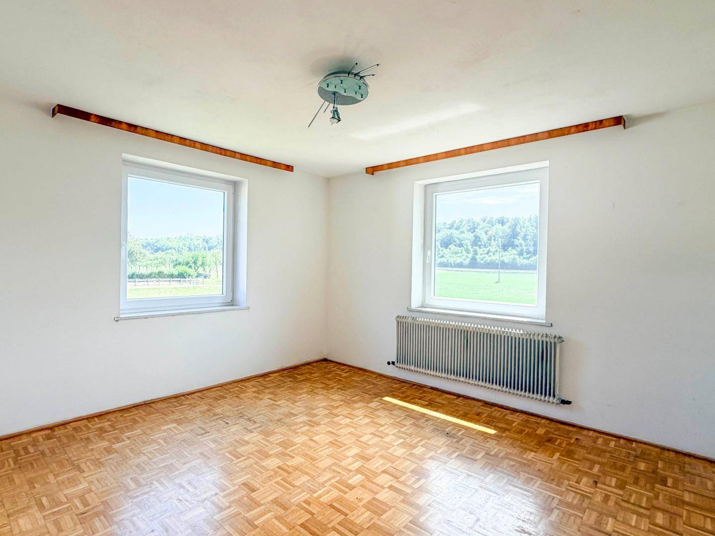 Heller Raum mit Parkettboden, Heizkörper und zwei Fenstern mit weitem Blick ins Grüne.