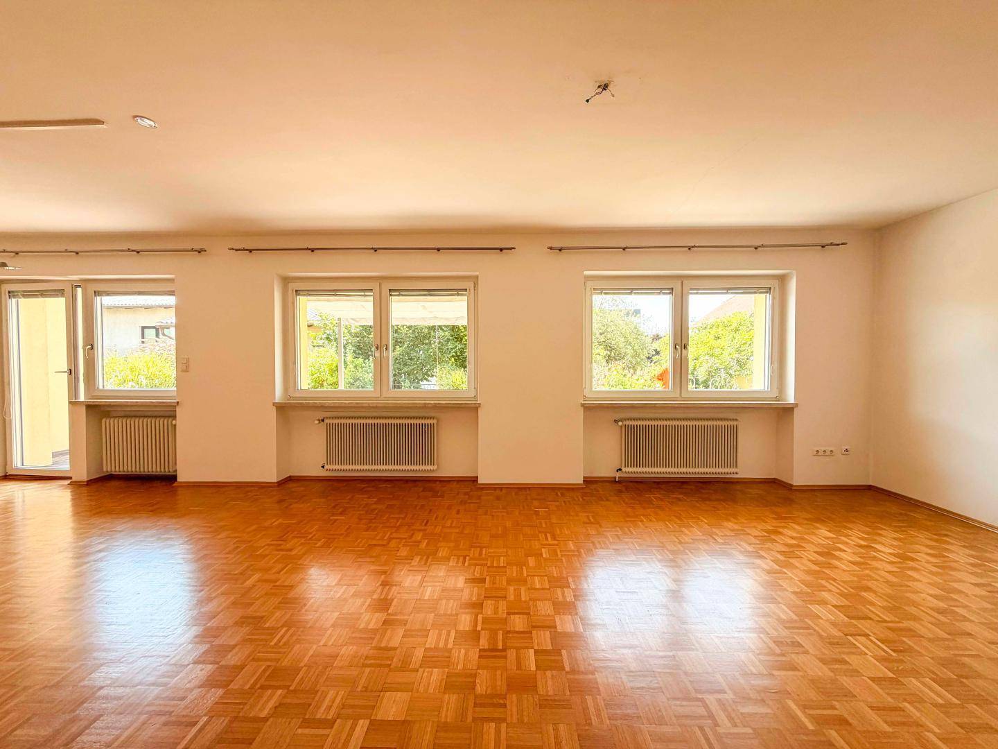 Helles Wohnzimmer mit Parkettboden und mehreren Fenstern mit Blick ins Grüne.