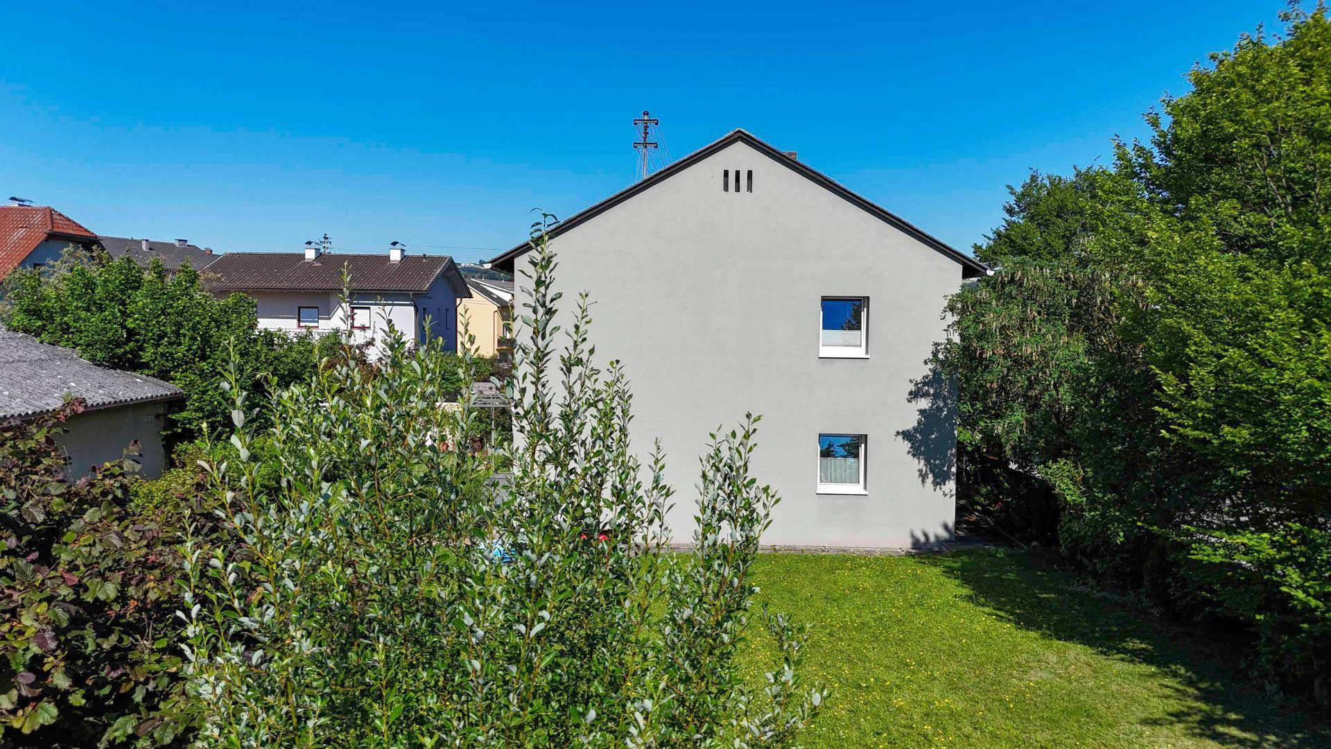 Rückansicht des Hauses mit schlichter Fassade und Fenstern, umgeben von Grün.