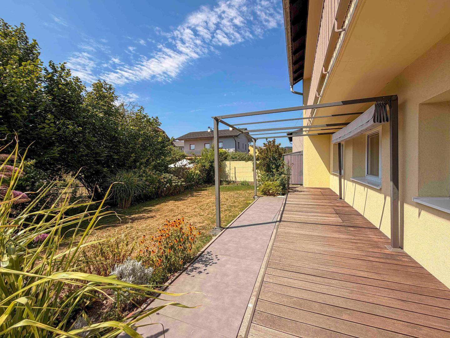 Lange Holzterrasse mit Pergola-Struktur und angrenzendem, sonnigen Gartenbereich.