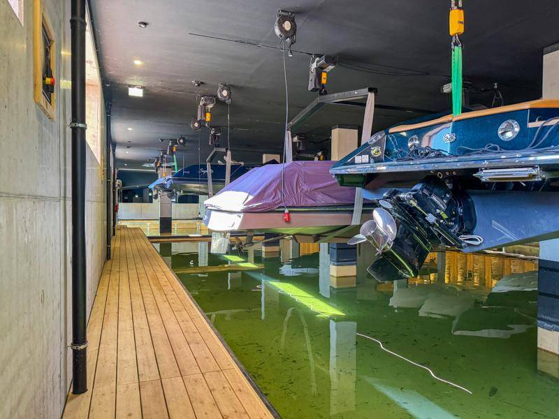 Innenansicht einer Bootsgarage mit Holzsteg und gelagerten Booten über dem Wasser.