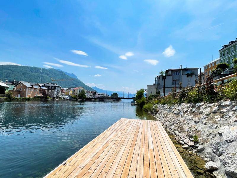 Langer Holzsteg am Flussufer mit Blick auf die Stadt und die umliegenden Berge.