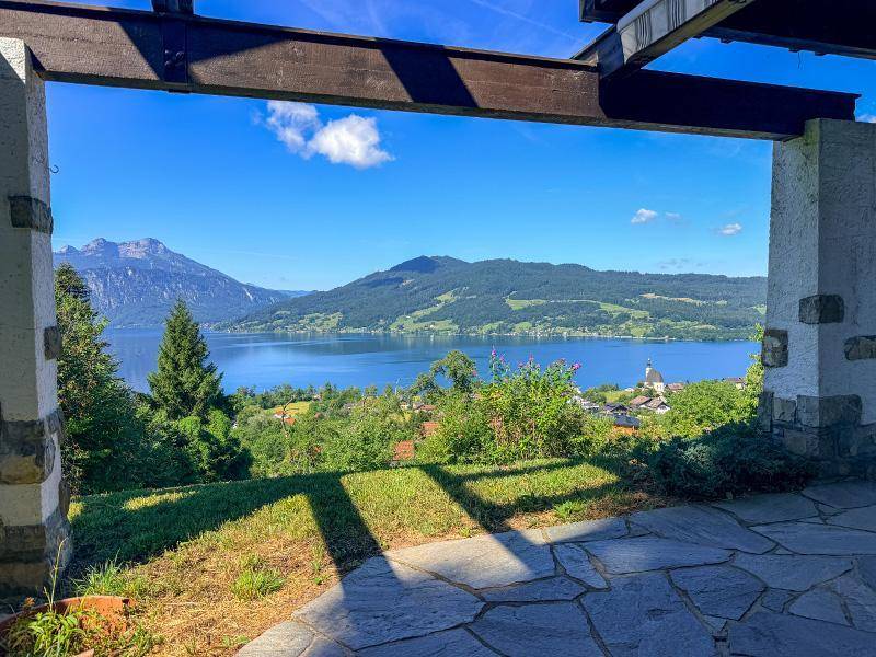 Überdachte Terrasse mit weitem Blick auf den See und die umliegende Berglandschaft unter blauem Himmel.