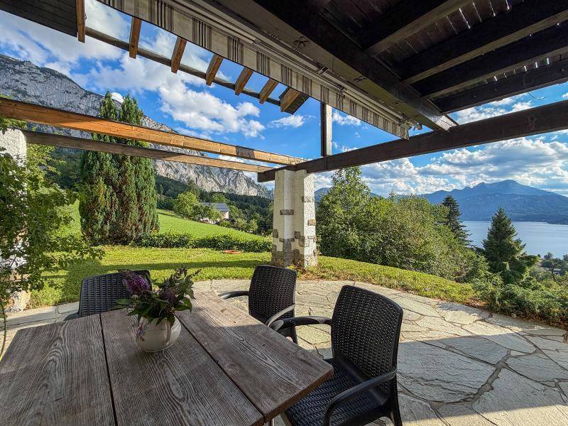 Möblierte Terrasse mit Holztisch und Stühlen, umgeben von Bergen und einem weiten Seeblick.