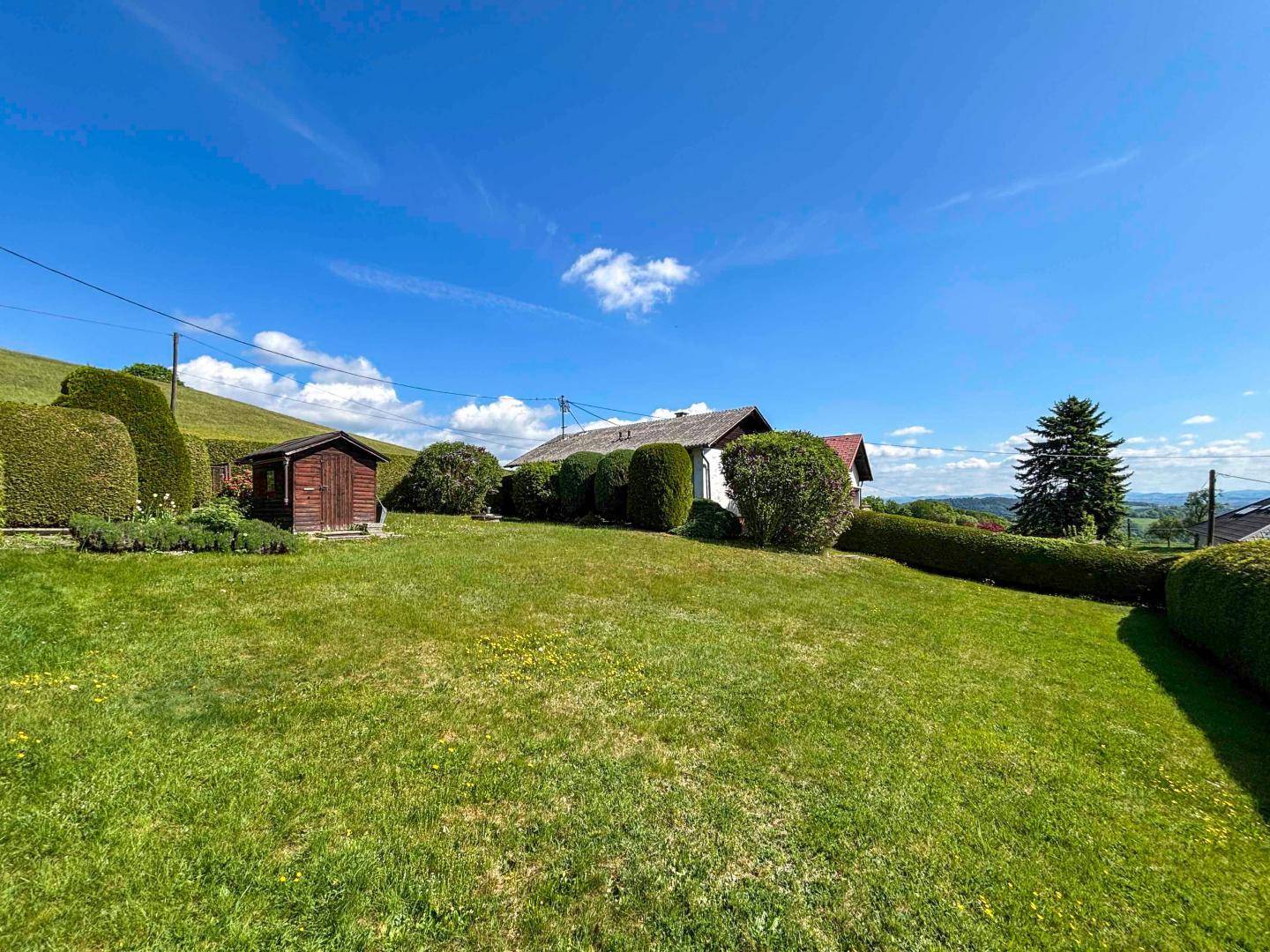 Weitläufiger Gartenbereich mit Rasen, Hecken und einem kleinen Schuppen vor hügeliger Landschaft.