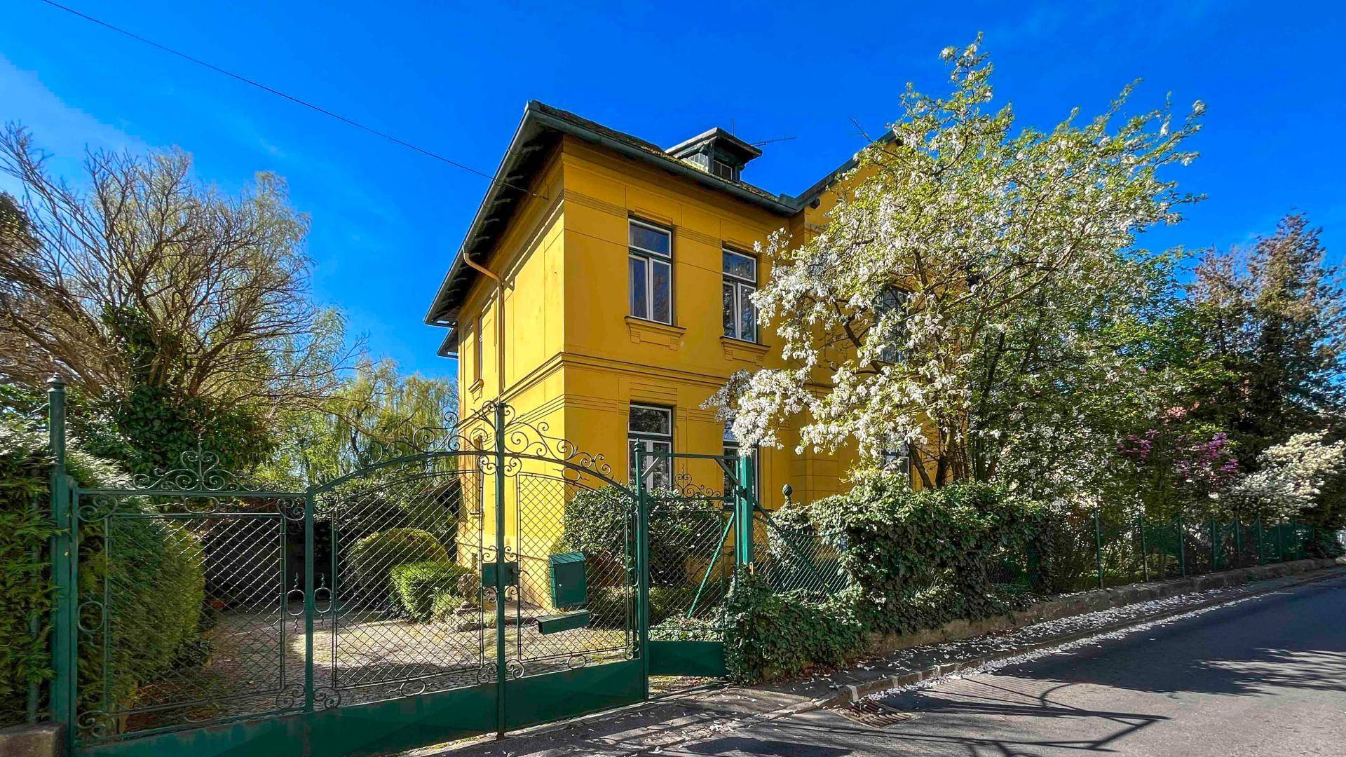 Historische Villa mit leuchtend gelber Fassade und schmiedeeisernem Tor in grüner Umgebung.
