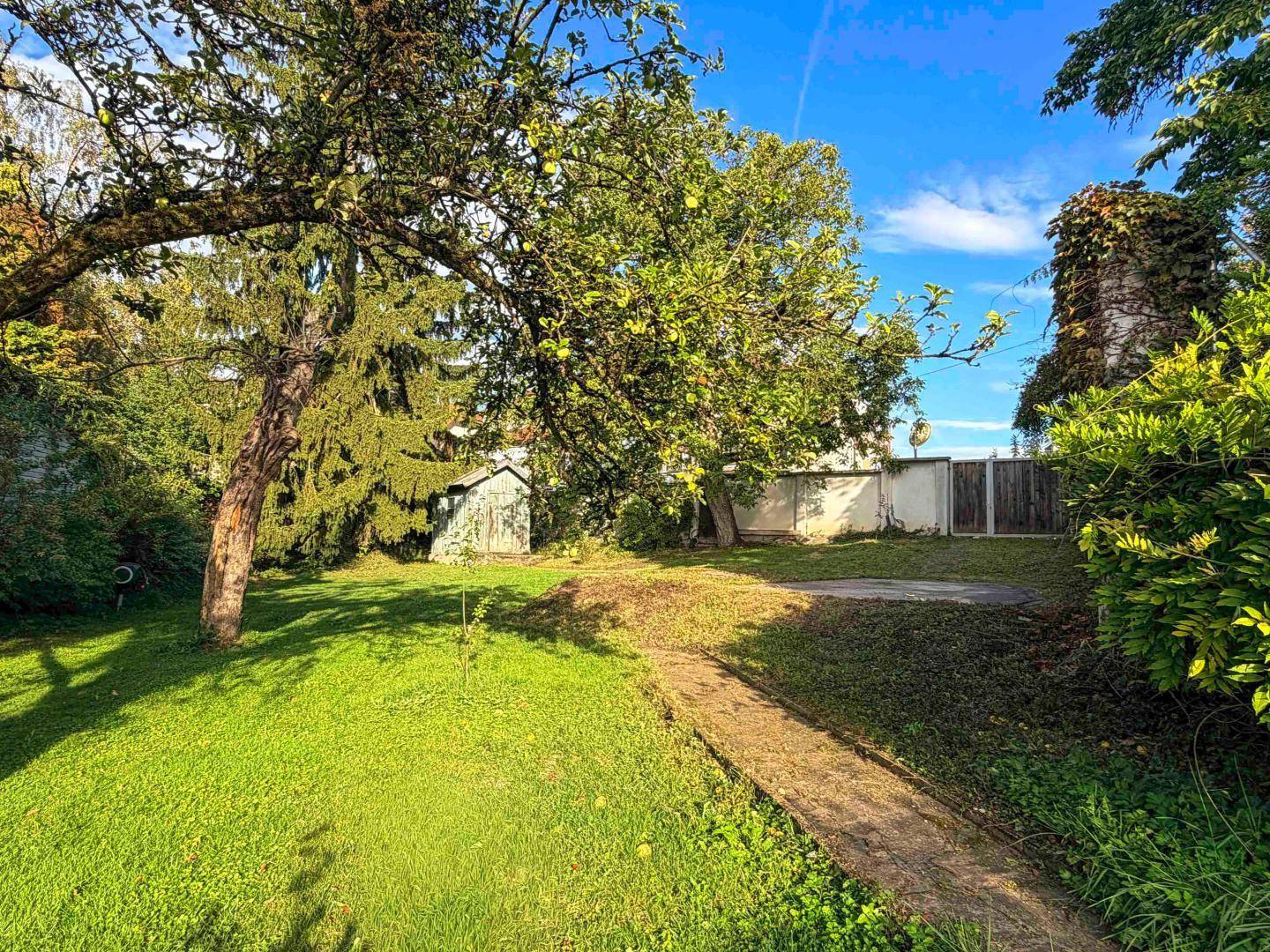 Weitläufiger Garten mit Rasen, altem Baumbestand und einem kleinen Gartenhaus unter blauem Himmel.