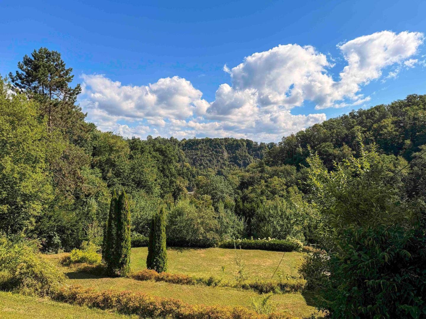 Weitläufige Gartenanlage mit Rasen und Bäumen, umgeben von bewaldeten Hügeln und Fernblick.