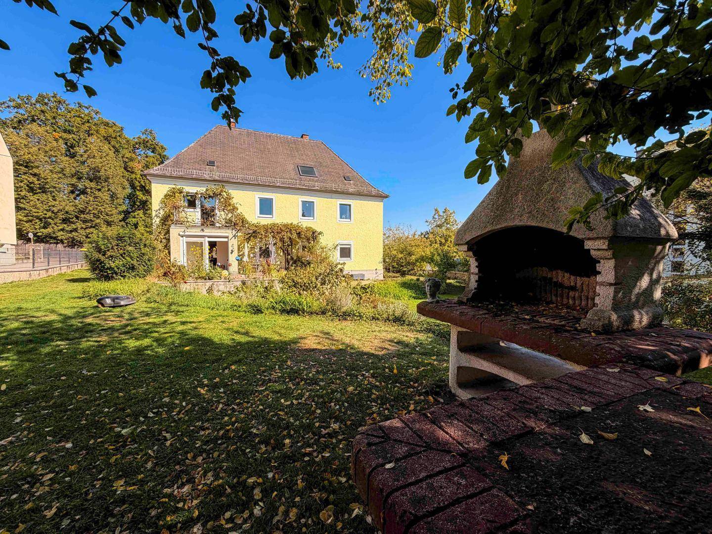 Gelbes Haus mit großem Garten und gemauertem Grillplatz unter blauem Himmel.