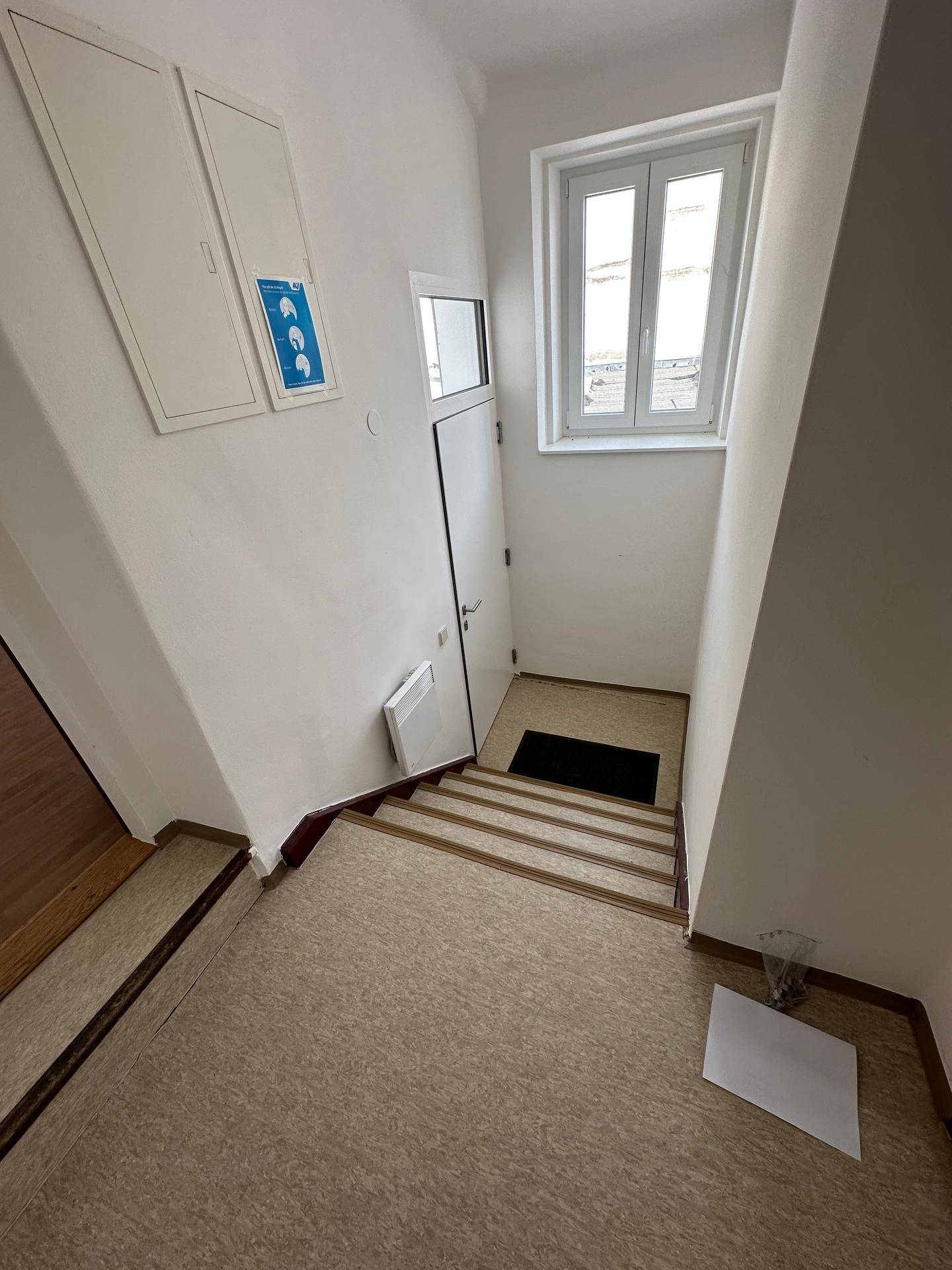 Heller Treppenaufgang mit Fenster, Linoleumboden und weißen Wänden, der zu den oberen Etagen führt.