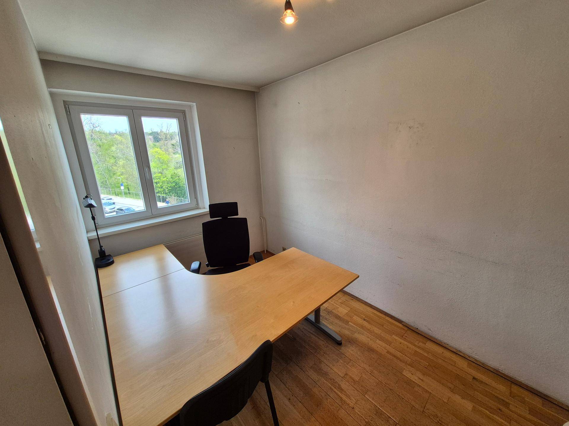 Helles Büro mit Holzboden, Schreibtisch und einem Fenster mit Blick ins Grüne.