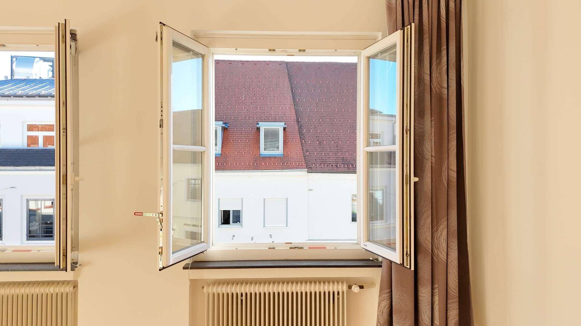 Geöffnetes Fenster mit Blick auf ein rotes Dach und angrenzende Gebäude, umgeben von beigen Wänden.