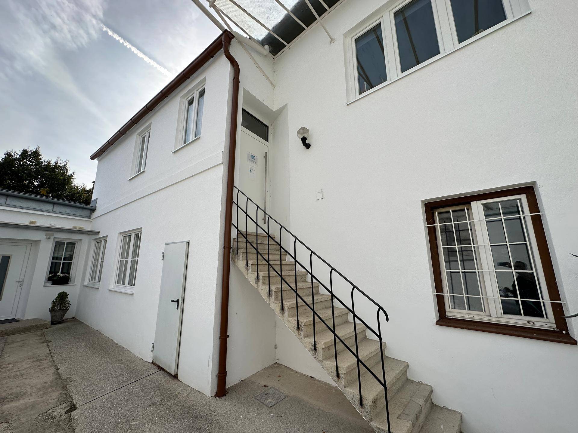 Weiß gestrichenes Gebäude mit Außentreppe und mehreren Fenstern unter einem bewölkten Himmel.