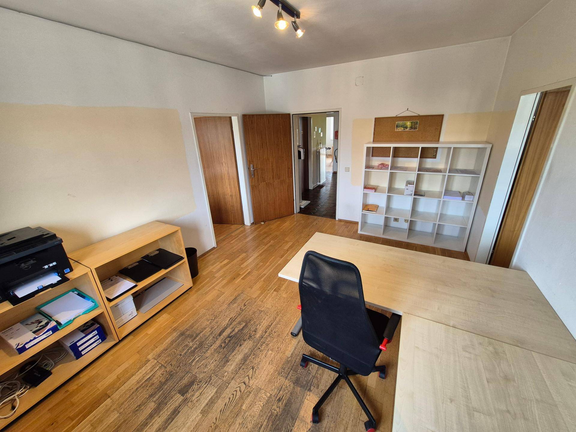 Büro mit Holzboden, Schreibtisch, Regalen und einem Durchgang zu einem weiteren Raum.