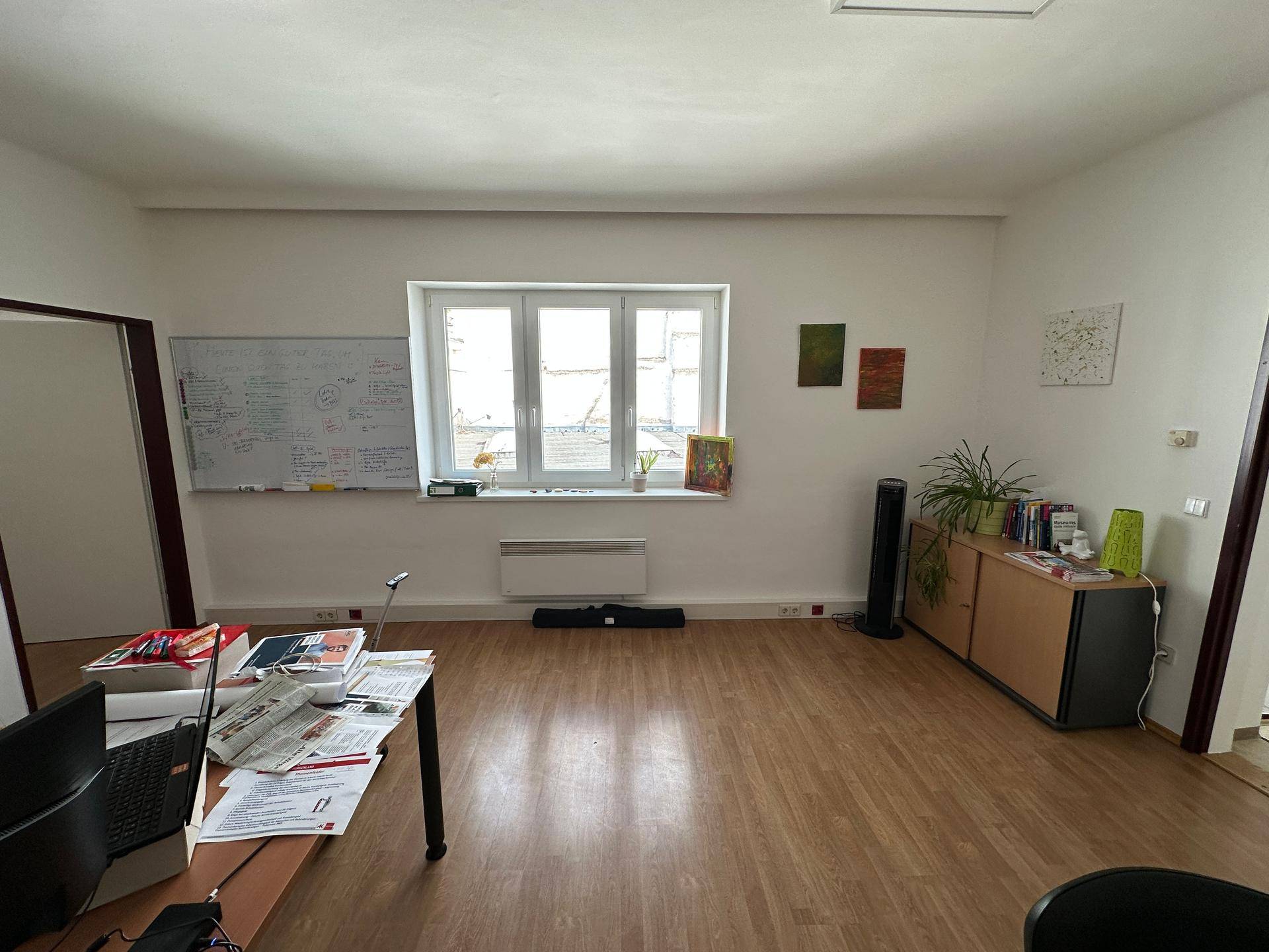 Helles Büro mit Holzfußboden, großem Fenster und einem Sideboard mit Pflanzen, das eine angenehme Atmosphäre schafft.
