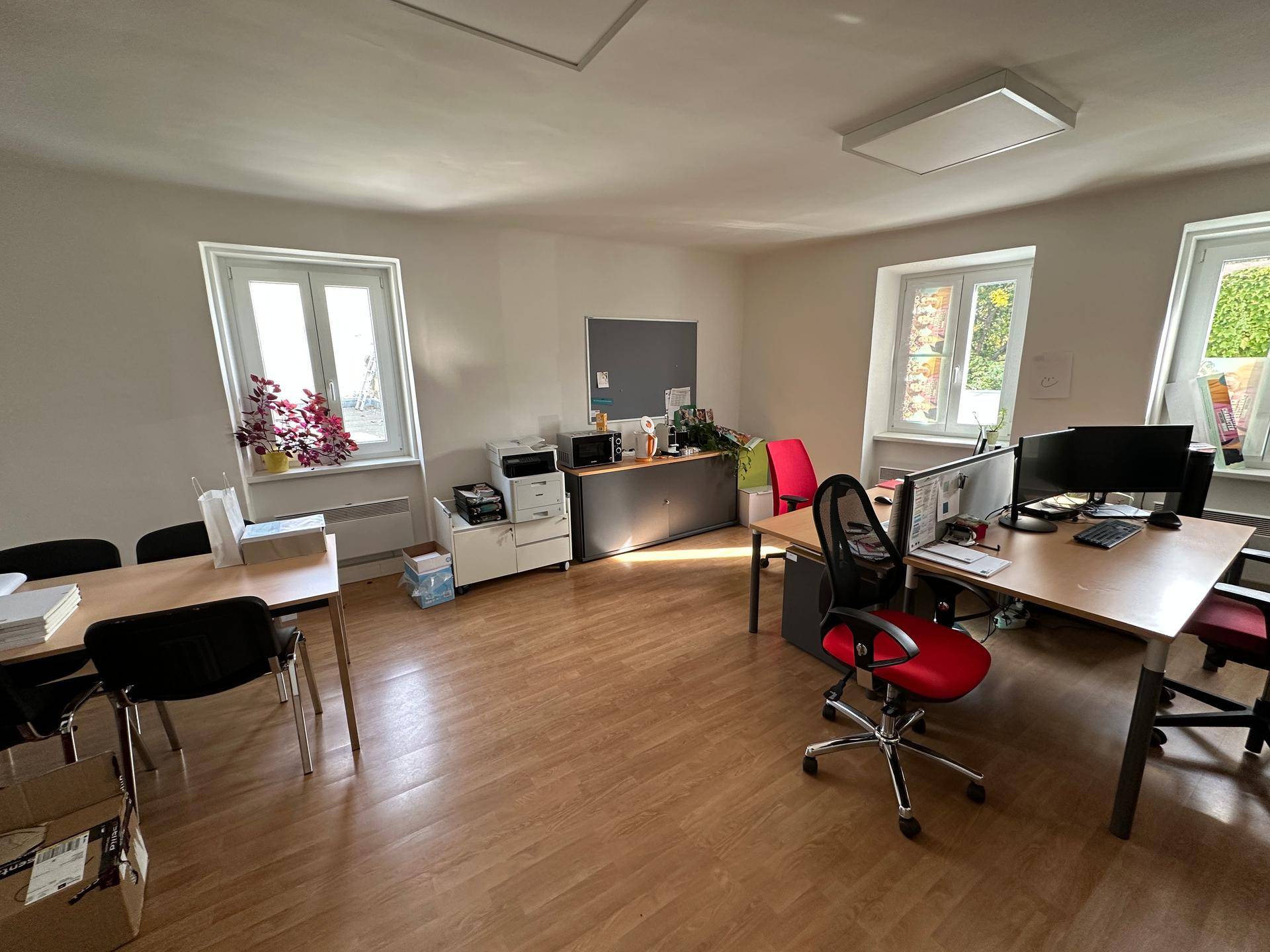 Geräumiges Büro mit mehreren Arbeitsplätzen, Holzfußboden und großen Fenstern, die viel Tageslicht hereinlassen.