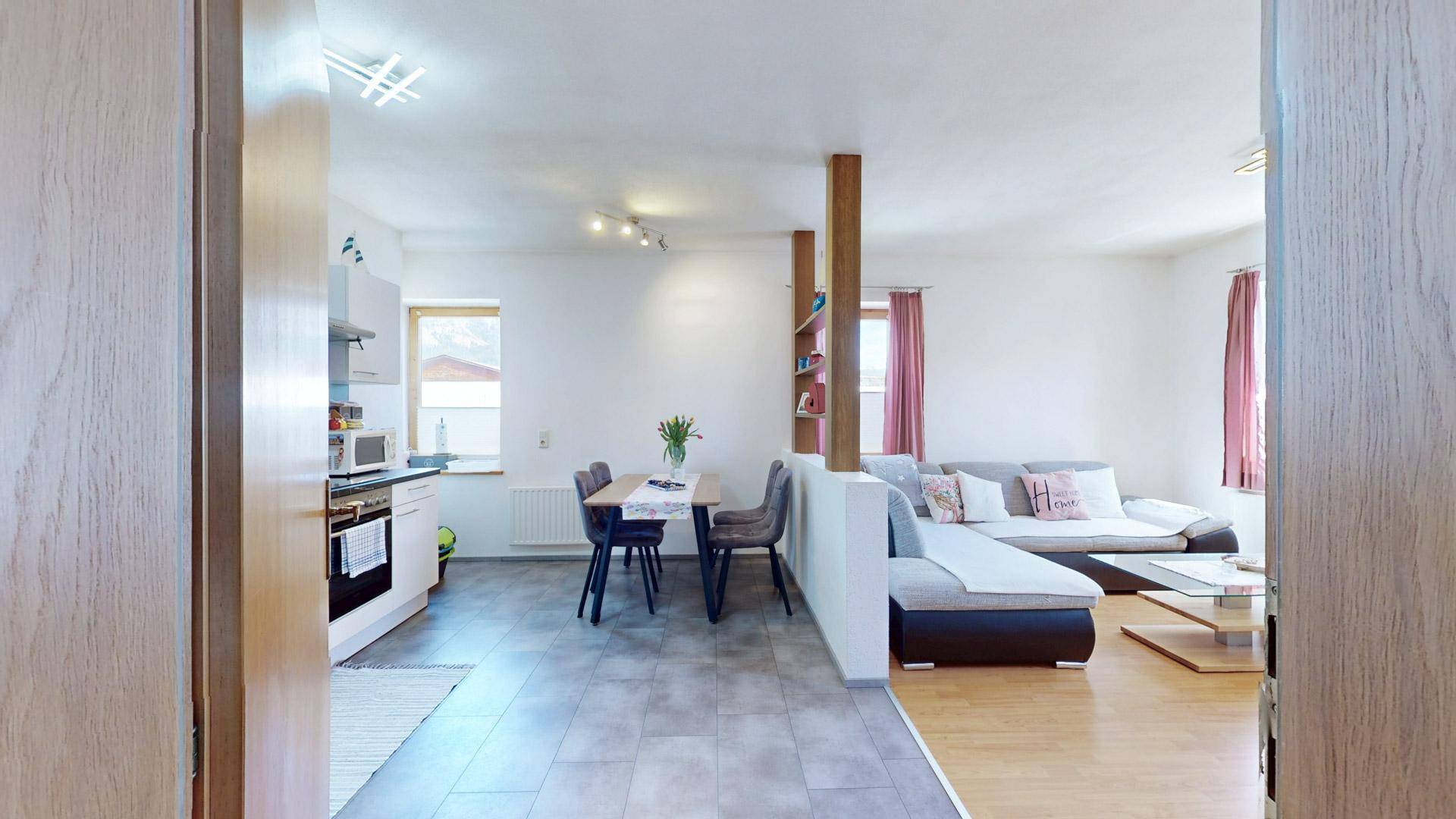 Offener Grundriss mit Blick von der Küche in den Wohnbereich mit Sofa und Fenstern.