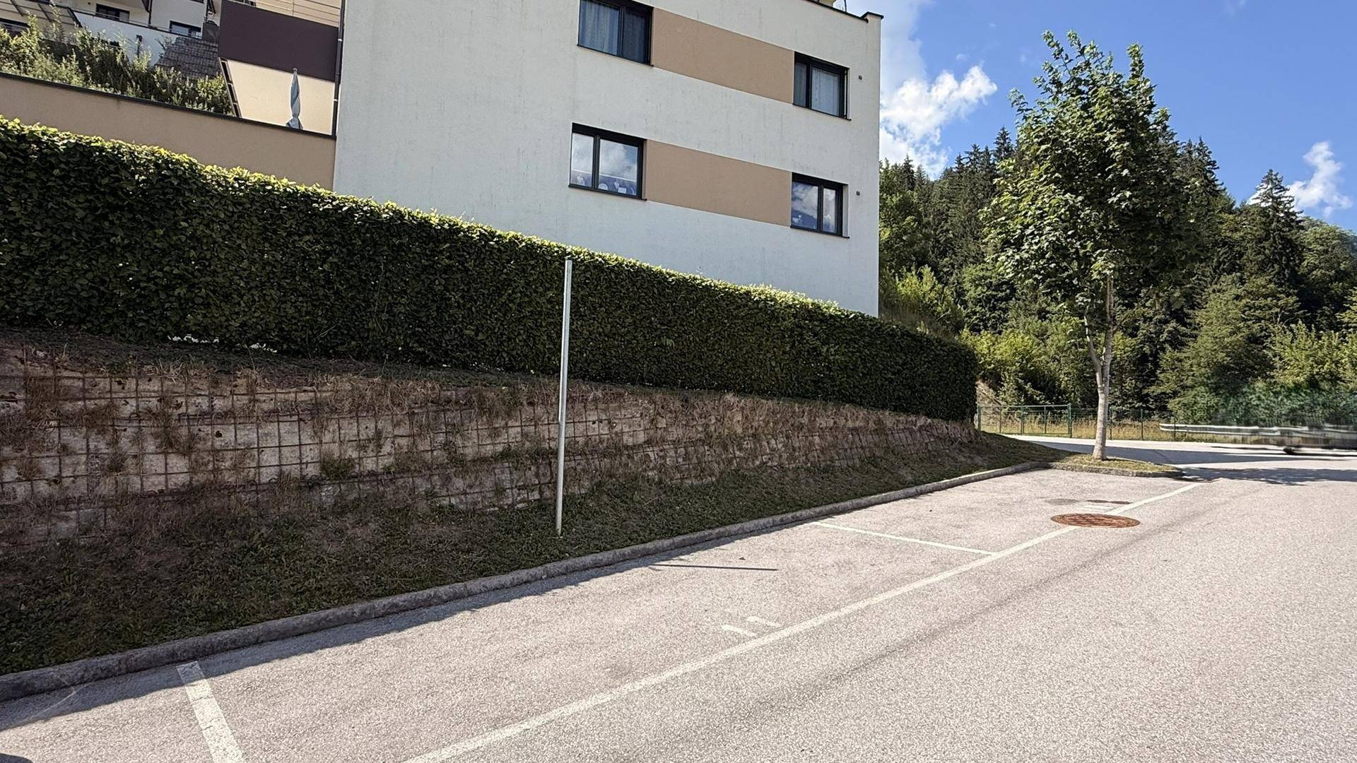 Mehrstöckiges Gebäude mit weiß-beiger Fassade, umgeben von einer hohen Hecke und Parkplätzen.