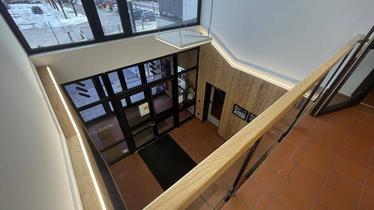 Blick von oben auf den Eingangsbereich mit Treppe, Glasfronten und Holzverkleidung, die viel Tageslicht hereinlassen.
