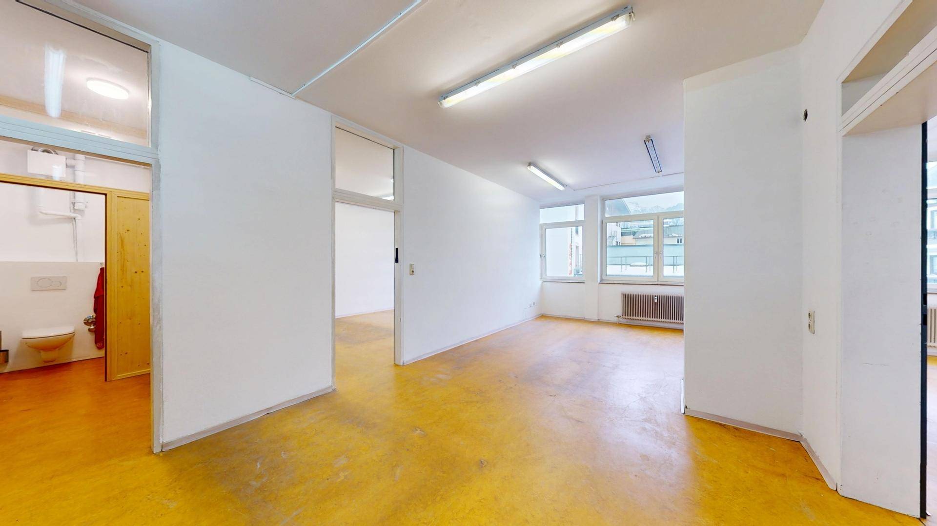 Geräumiger Bürobereich mit gelbem Boden, weißen Wänden und Zugang zu einer Toilette.
