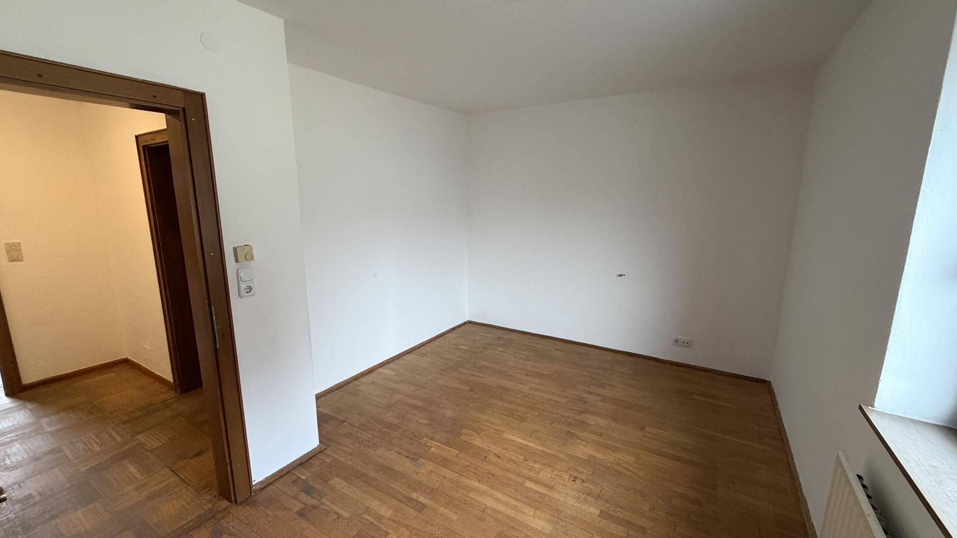 Leeres Zimmer mit Parkettboden und weiß gestrichenen Wänden, bereit zur Gestaltung.